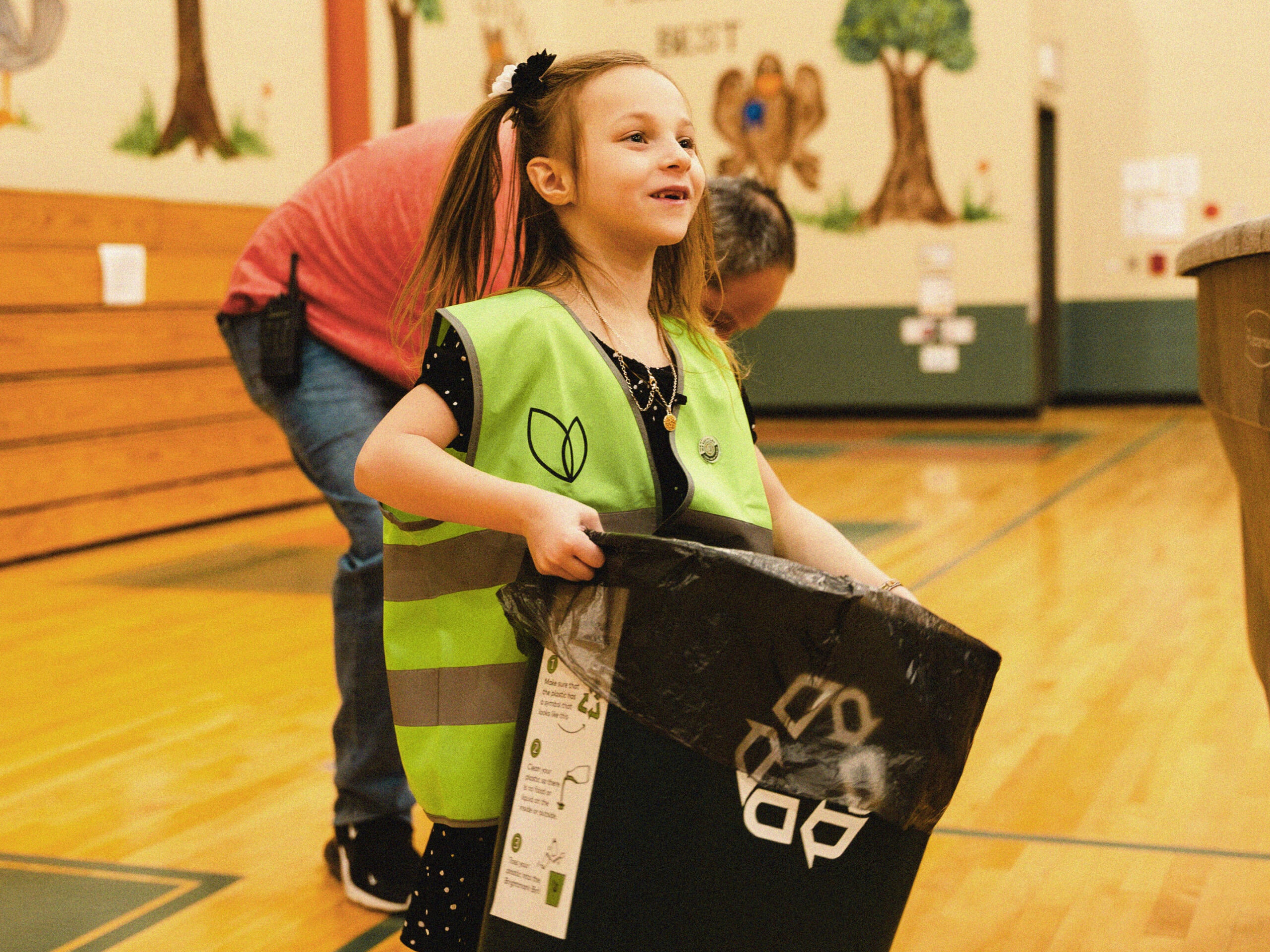 Young girl wearing a Brightmark safety vest and carrying a recycling bin.